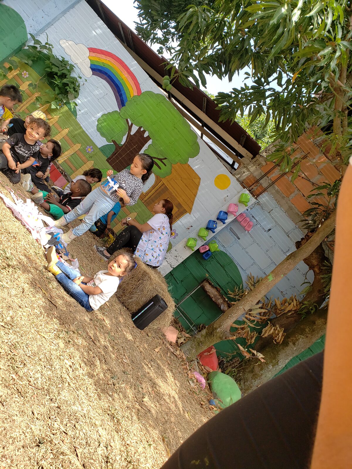 Niñas y niños sentados en el suelo del patio, junto a un muro con un mural de árboles y un arcoíris