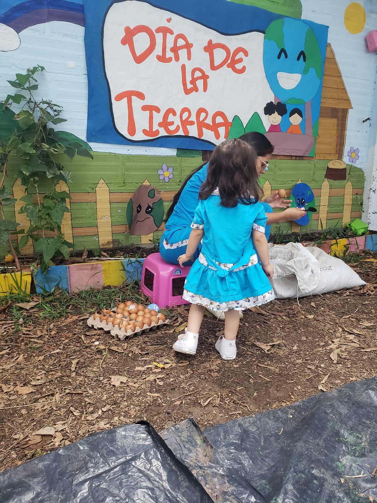 Niña de vestido azul y una docente junto al mural del Día de la Tierra