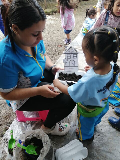 Docente de uniforme azul ayuda a una niña de trenzas a llenar de tierra una bandeja de siembra en el patio