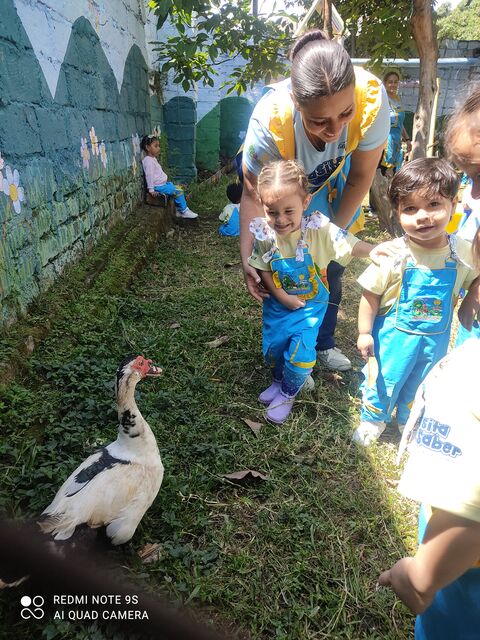 Docente inclinada junto a dos niños de overol azul que caminan sobre el césped, al lado de un muro con mural de montañas