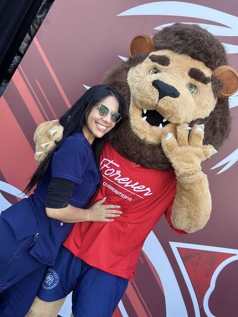 Mujer con uniforme azul y gafas de sol posa abrazada a una mascota de león de peluche gigante con camiseta roja, frente a un mural rojo y blanco.