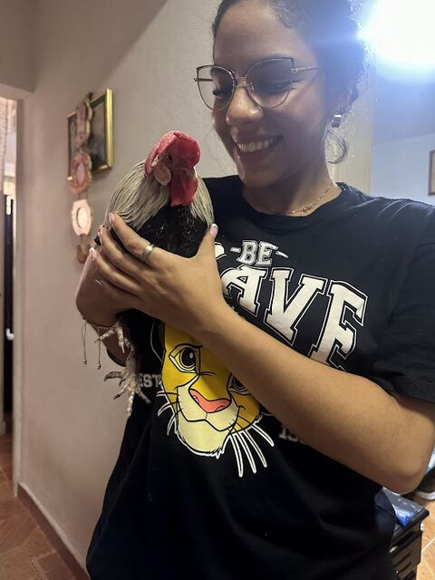 Mujer joven con gafas y camiseta negra estampada sostiene en brazos un gallo de cresta roja, sonriendo, dentro de una casa.