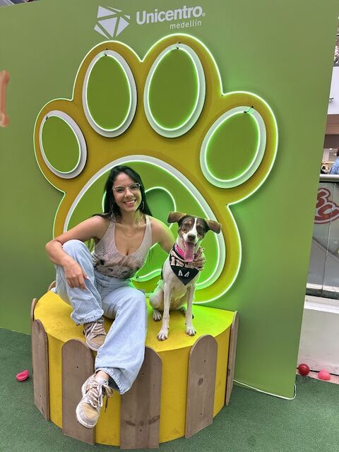 Mujer sentada junto a un perro pequeño blanco y café sobre una tarima amarilla, frente a una gran huella verde decorativa con luces de neón en un centro comercial.