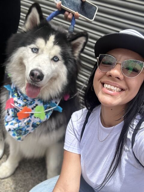 Mujer sonriente con gorra y gafas se toma una selfie junto a un perro husky que lleva una pañoleta de colores