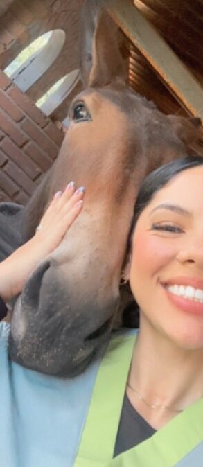 Selfie de una mujer sonriente con uniforme azul claro que acaricia y apoya la cabeza contra el hocico de un caballo café en una pesebrera.