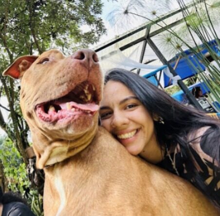 Selfie al aire libre de una mujer sonriente junto a un perro grande tipo pitbull de pelaje café claro con la boca abierta, entre árboles.
