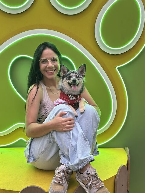 Mujer con gafas sentada con las piernas recogidas sostiene en su regazo a un perro pequeño de pelaje gris con pañoleta roja, frente a una huella verde gigante iluminada con neón.