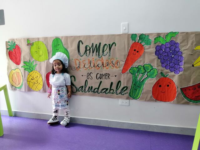 Niña con gorro de chef frente a un mural de frutas y verduras que dice «Comer delicioso es comer saludable»
