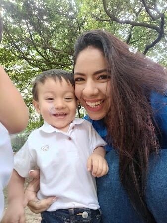 Selfie de una mujer sonriente abrazando a un niño pequeño de camisa blanca, al aire libre bajo árboles
