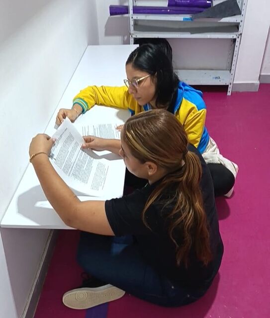 Dos mujeres sentadas en el piso revisando y diligenciando documentos sobre una mesa blanca durante una jornada de capacitación