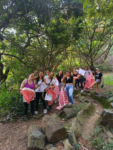 Grupo numeroso de mujeres posando al aire libre bajo árboles frondosos, con manteles de cuadros rojos y hojas en la mano, durante una jornada de formación