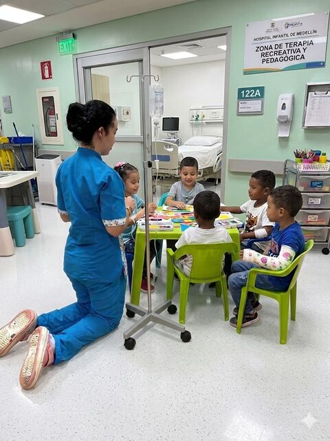Docente de uniforme azul arrodillada junto a una mesa verde donde varios niños pequeños realizan una actividad, en una zona de terapia recreativa y pedagógica de un hospital infantil