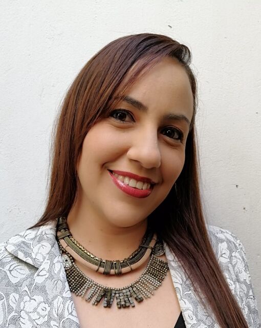 Retrato de Fabiola, cofundadora: cabello castaño largo, sonriente, con collar artesanal y chaqueta clara sobre fondo blanco.