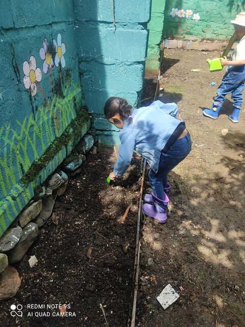 Niña con chaqueta de jean y botas moradas sembrando en la huerta junto a un muro pintado con flores, mientras otro niño riega al fondo
