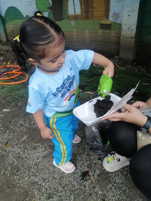 Niña pequeña con camiseta de Mi Casita del Saber echando tierra con una pala verde en una bandeja durante una actividad de siembra