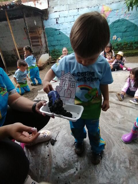 Niño con camiseta de Mi Casita del Saber, cabeza agachada, echando tierra con una pala azul en una bandeja que sostiene una docente, frente a un muro pintado con montañas y flores