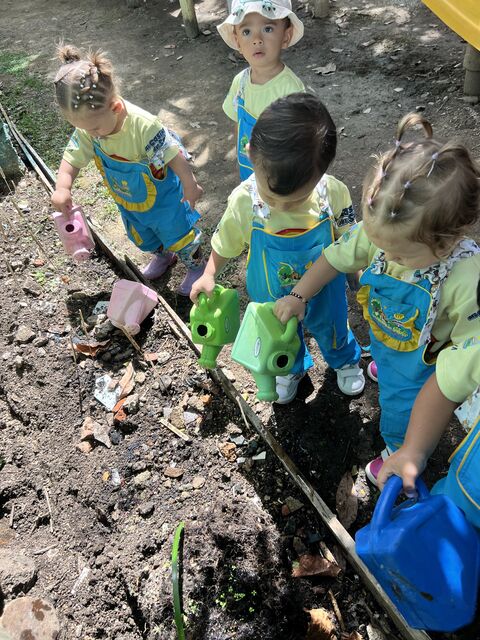 Cuatro niños con delantal azul y camiseta amarilla regando la huerta con regaderas de colores