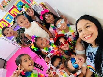 Docente tomándose una selfie con un grupo de niños con collares hawaianos, coronas de flores y gafas de colores