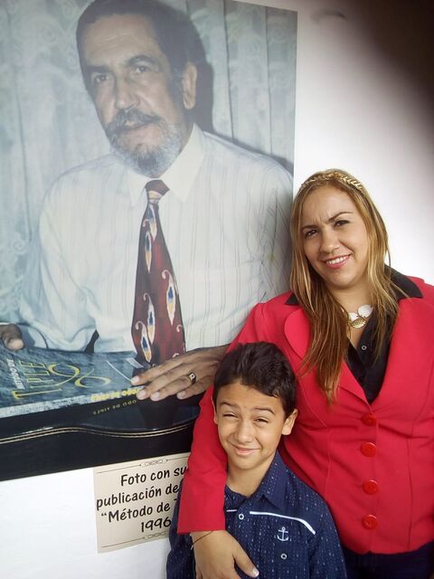 Una mujer de saco rojo posa junto a un retrato de gran formato del Maestro Elkin Pérez Álvarez, de camisa blanca y corbata roja