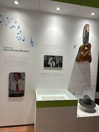 Exposición en homenaje al Maestro Elkin Pérez Álvarez: muro blanco con pentagramas azules, un instrumento de cuerda exhibido en la pared, una vitrina con una partitura y un sombrero gris