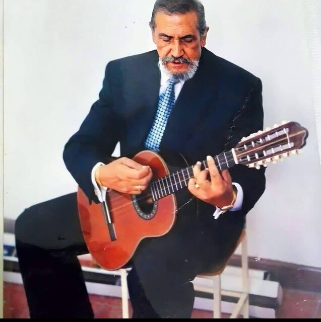 El Maestro Elkin Pérez Álvarez sentado, de traje oscuro y corbata azul, tocando un instrumento de cuerda