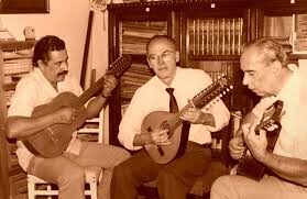 Fotografía antigua en tono sepia de tres músicos sentados tocando instrumentos de cuerda —guitarra, bandola y tiple— frente a una biblioteca