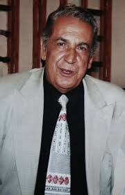 Retrato del Maestro Elkin Pérez Álvarez en edad madura, con saco claro, camisa negra y corbata bordada