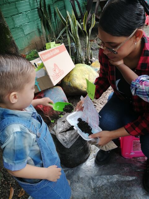 Maestra de camisa a cuadros y un niño pequeño con overol de jean preparando tierra y semillas en una actividad de siembra.