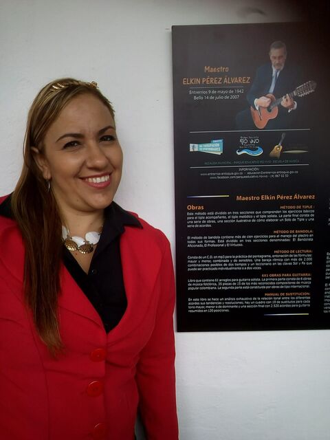 Mujer sonriente de chaqueta roja junto al pendón conmemorativo del Maestro Elkin Pérez Álvarez, con su retrato tocando guitarra y la reseña de sus obras.