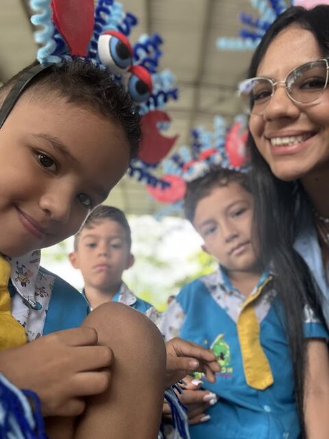 Selfie de una maestra con gafas, sonriente, junto a niños con uniforme azul y diademas de colores hechas a mano.