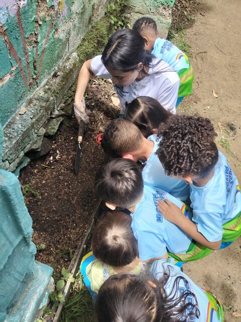 Grupo de niños con uniforme azul y su maestra, agachados sembrando en la tierra de la huerta.